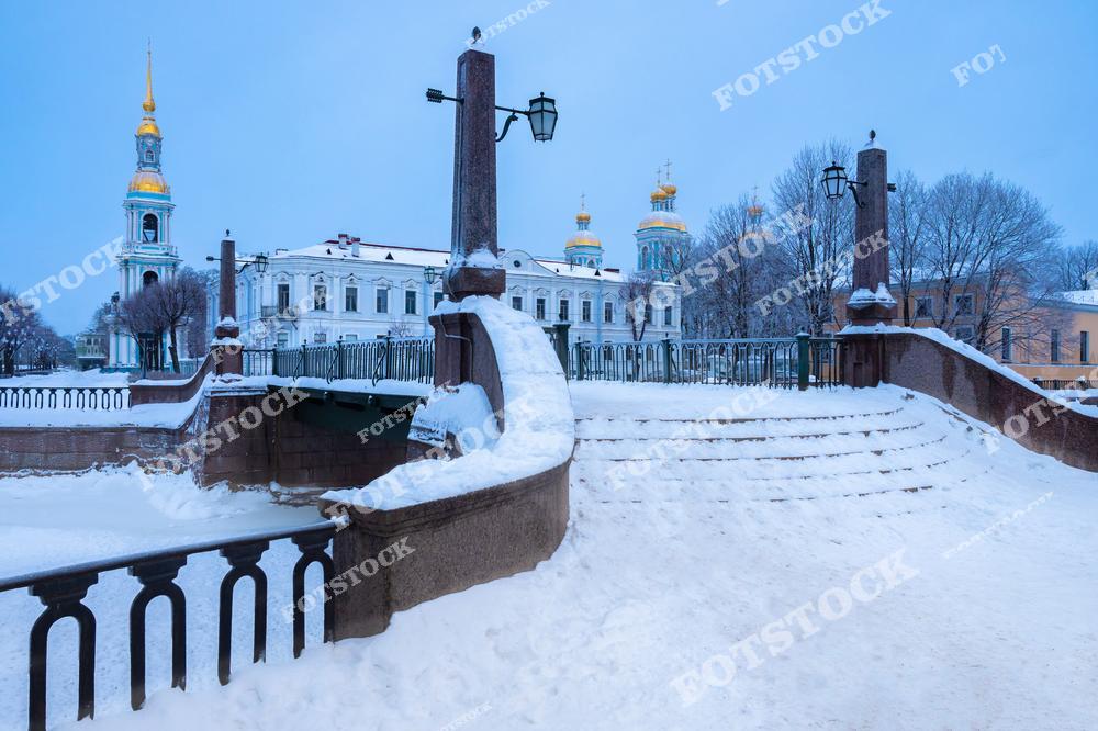 На переднем плане изображены ступени, ведущие к набережной канала, покрытые пушистым снегом. Вдали возвышается величественный Никольский морской собор с его золотыми куполами, контрастирующими с голубоватым утренним небом. Архитектура моста и фонари придают этому виду атмосферу уюта и спокойствия. Этот зимний пейзаж отражает неповторимую красоту Санкт-Петербурга.
